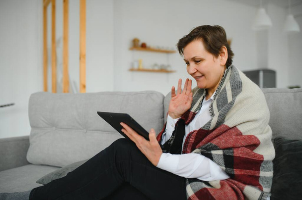 Middle-aged woman reading a message, e-book or information on her tablet computer with a look of excited anticipation as she sits on a couch at home - Zdjęcie, obraz