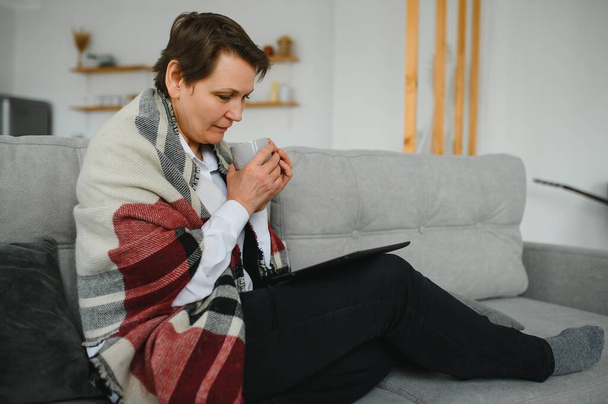 Keski-ikäinen nainen lukee viestiä, e-kirjaa tai tietoa tablet-tietokoneellaan innoissaan odotuksesta, kun hän istuu sohvalla kotona.
 - Valokuva, kuva
