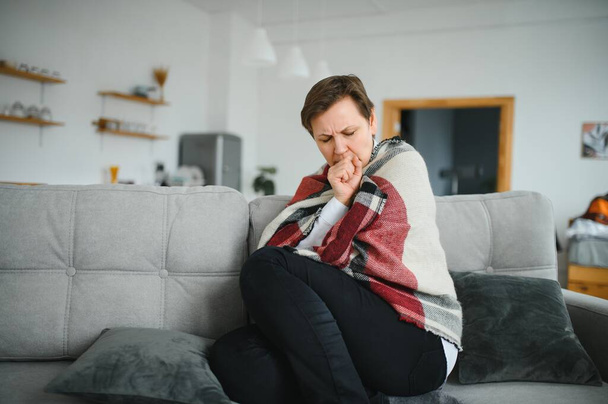 Middle-aged 50s sick frozen woman seated on sofa in living room covered with warm plaid sneezing, runny nose feels unhealthy, seasonal cold, weakened immune system concept - 写真・画像