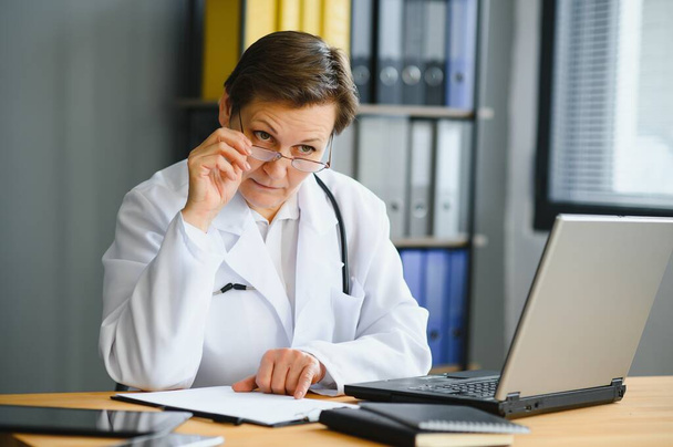 Travailleuse de la santé souriante faisant de la paperasse et utilisant un ordinateur portable tout en travaillant au bureau du médecin. - Photo, image