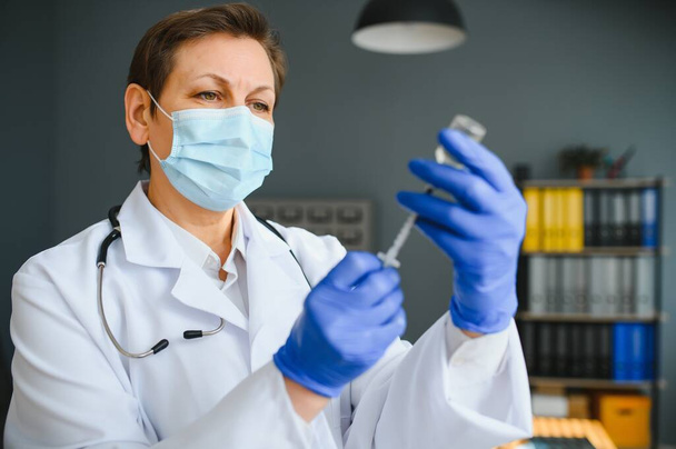 Zbliżenie ręki lekarza lub naukowca w mundurze lekarza noszącego maskę ochronną w laboratorium zawierającym szczepionkę zawierającą płyn w butelce, koronawirus lub koncepcję COVID-19. - Zdjęcie, obraz