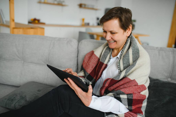 portrait of beautiful old woman using tablet pc and smiling, sitting on sofa at home - Фото, изображение