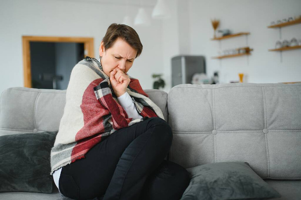 Middle-aged 50s sick frozen woman seated on sofa in living room covered with warm plaid sneezing, runny nose feels unhealthy, seasonal cold, weakened immune system concept - Fotografie, Obrázek