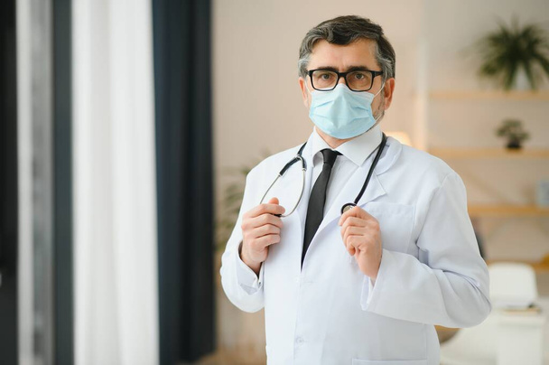 Mature old medical healthcare professional doctor wearing white coat, stethoscope, glasses and face mask. Medical staff health care protection concept. Portrait - Photo, Image