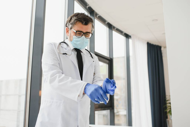 Professional doctor in medical mask putting on gloves. Confident physician in lab coat wearing protection during covid-19 outbreak. Prevention of viral diseases - Foto, immagini