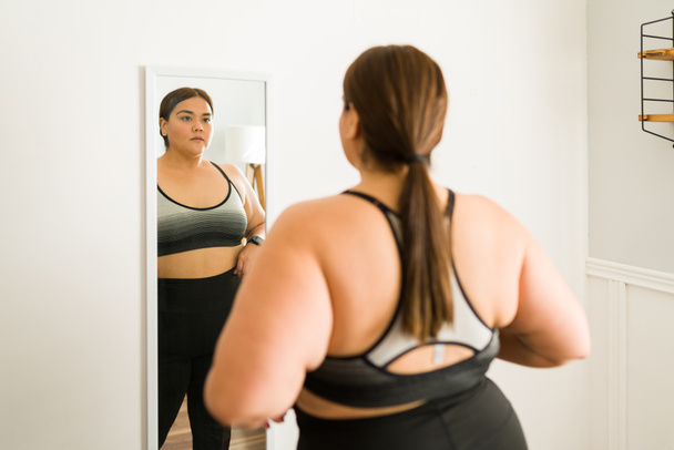 Afvallen voor mijn gezondheid. Bezorgde jonge vrouw met obesitas dieet en kijken zichzelf in de spiegel - Foto, afbeelding