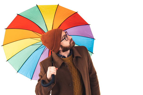 Portrait of young handsome bearded Caucasian man holding a multicoloured umbrella and looking up at the sky weather forecast concept brown casual attire isolated white background studio shot  - 写真・画像