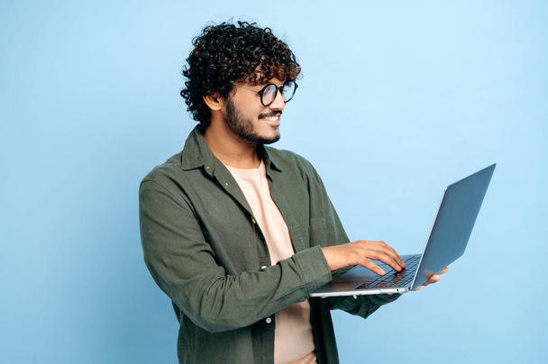 Умный современный индийский или арабский парень в очках, стильно одетый, держит в руках ноутбук, просматривает информацию, интернет, чаты в социальных сетях, стоит на изолированном синем фоне, улыбается - Фото, изображение