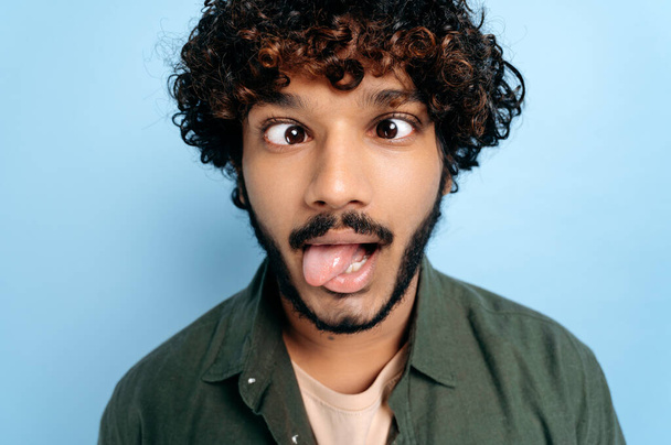 Close up face of indian or arabian curly funny guy, making a funny face, tongue out, squinted his eyes, standing on isolated blue background. Grimaces, fooling around - Fotoğraf, Görsel