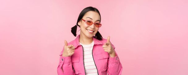 Крупный план портрет современной азиатской девушки в солнечных очках улыбается, указывая пальцами на камеру, хвалить вас, приглашая или комплимент, стоя на розовом фоне - Фото, изображение