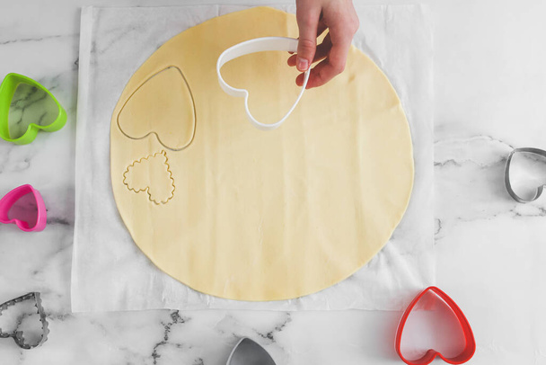 Hands of caucasian teenage girl holding plastic heart cutter over round dough on white parchment . - Photo, Image
