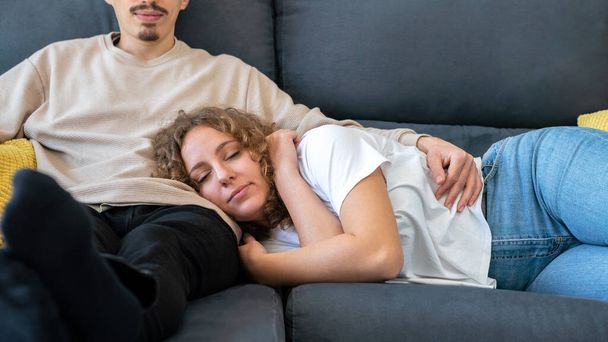 Couple relaxing in sofa at cozy apartment. Sleeping woman with curly hair taking nap on the sofa while boyfriend watching TV. Attractive girl sleep lying on the comfortable couch with eyes closed - Photo, Image