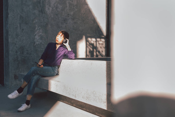 Feliz joven asiática mujer escuchando música con auriculares a través de teléfono inteligente y divertirse mientras se sienta al lado de la calle. - Foto, imagen