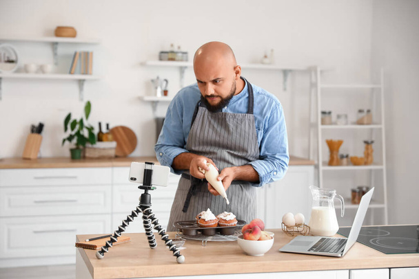 Bell'uomo che decora i muffin alla pesca con la crema mentre segue il video tutorial in cucina - Foto, immagini