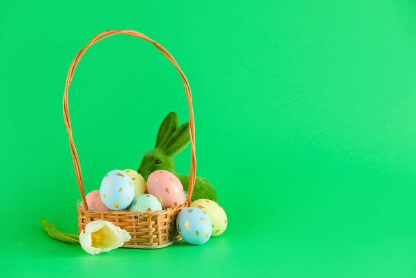 Wicker basket of painted Easter eggs, flower and toy bunny on green background - Photo, Image