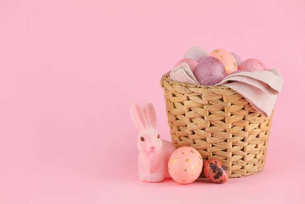 ピンクの背景にイースターエッグとおもちゃのウサギを描いたウィッカーバスケット - 写真・画像