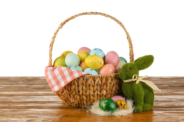 Плетений кошик з розфарбованих великодніх яєць та іграшкового кролика на столі на білому тлі
 - Фото, зображення