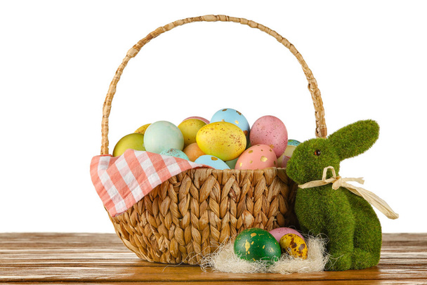 Cesta de vime de ovos de Páscoa pintados e coelho de brinquedo na mesa contra fundo branco - Foto, Imagem