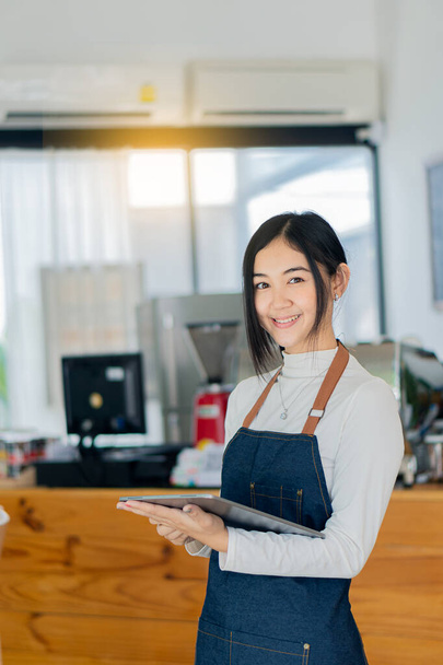 A beautiful Asian barista in an apron holds a platelet and stands in front of a coffee shop counter. Attractive start-up ideas for business owners. - Photo, Image