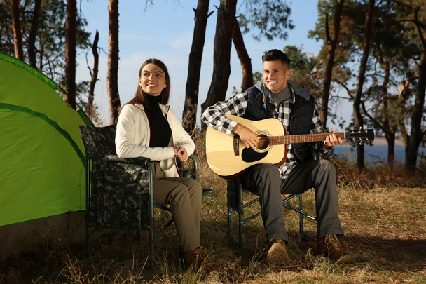 Couple with guitar resting in camping chairs near tent outdoors - Foto, afbeelding