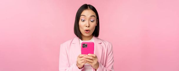 Retrato de mujer de negocios asiática con la cara sorprendida, usando aplicación de teléfono inteligente, usando traje de negocios. Chica coreana con teléfono móvil y expresión de la cara emocionada, fondo rosa - Foto, Imagen