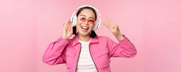 Dancing stylish asian girl listening music in headphones, posing against pink background - Фото, изображение