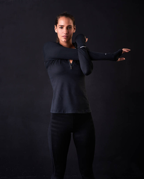 Focused on fitness. Studio portrait of a young woman in gym clothes stretching her arms against a dark background. - Фото, изображение