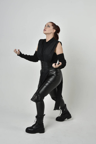 Volledig portret van mooie roodharige vrouwelijke model dragen zwarte futuristische scifi lederen kostuum. Dynamische staande poses met gebaren handen op een witte studio achtergrond. - Foto, afbeelding
