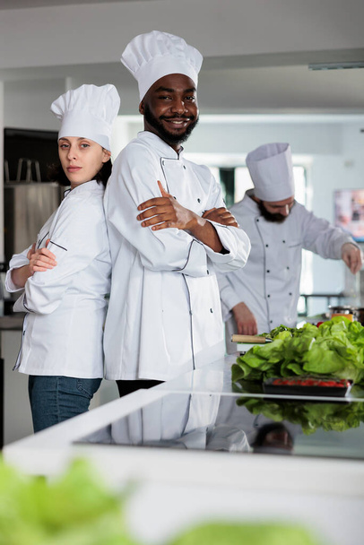 Multiethnische Gastro-Experten stehen mit verschränkten Armen in der Restaurantküche und posieren vor der Kamera. - Foto, Bild