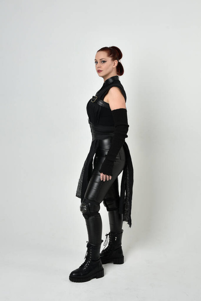 Full length portrait of pretty red haired female model wearing black futuristic scifi leather costume. Dynamic standing poses with gestural hands on a white studio background. - Foto, immagini