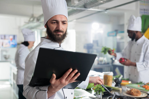 Food industry worker having laptop, following gourmet dish recipe on screen stirring ingredients in pan - Fotó, kép