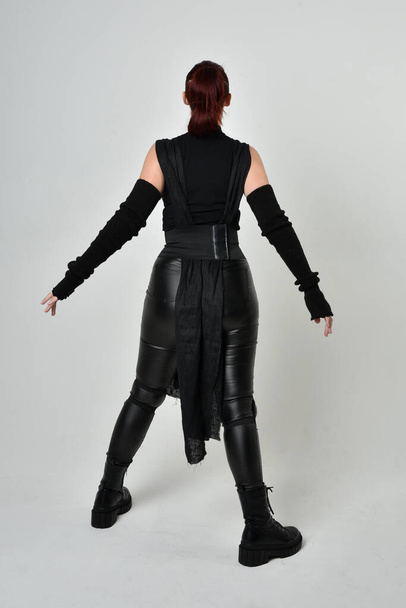 Volledig portret van mooie roodharige vrouwelijke model dragen zwarte futuristische scifi lederen kostuum. Dynamische staande houdingen met gebaren handen, naar achteren gericht weg van op een witte studio achtergrond. - Foto, afbeelding