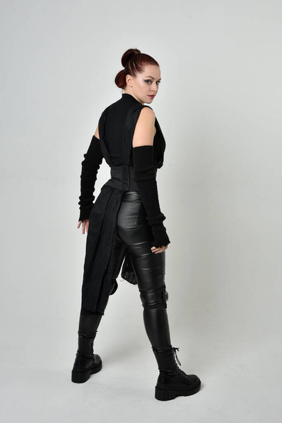Retrato de comprimento total do modelo feminino ruivo bonito vestindo traje de couro scifi futurista preto. Dynamic em pé posa com as mãos gestuais, viradas para trás de um fundo de estúdio branco. - Foto, Imagem