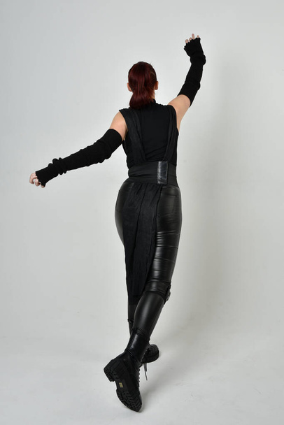 Full length portrait of pretty red haired female model wearing black futuristic scifi leather costume. Dynamic standing poses with gestural hands, facing backwards away from  on a white studio background. - Foto, Bild