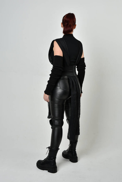 Full length portrait of pretty red haired female model wearing black futuristic scifi leather costume. Dynamic standing poses with gestural hands, facing backwards away from  on a white studio background. - Foto, imagen