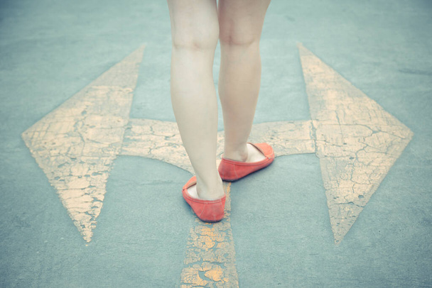 Vintage τόνος του κοριτσιού φορούν κόκκινα παπούτσια με τα πόδια προς την κατεύθυνση με κίτρινο σήμα βέλος κυκλοφορίας σε ένα ασφαλτοστρωμένο δρόμο φόντο - Φωτογραφία, εικόνα