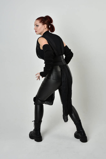 Portrait complet de jolie femme aux cheveux roux portant un costume futuriste noir en cuir de scifi. poses debout dynamiques avec des mains gestuelles, face à l'arrière sur fond de studio blanc. - Photo, image