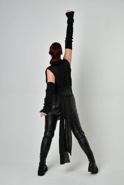 Retrato de comprimento total do modelo feminino ruivo bonito vestindo traje de couro scifi futurista preto. Dynamic em pé posa com as mãos gestuais, viradas para trás de um fundo de estúdio branco. - Foto, Imagem