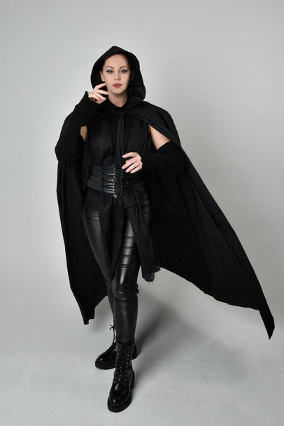 Retrato de comprimento total do modelo feminino ruivo bonito vestindo traje de couro scifi futurista preto com capa de fluxo preto camuflado. Dynamic em pé posa com as mãos gestuais, viradas para trás de um fundo de estúdio branco. - Foto, Imagem