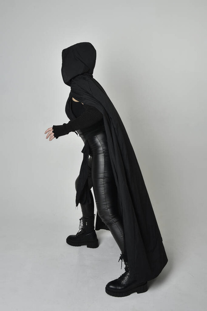 Full length portrait of pretty red haired female model wearing black futuristic scifi leather costume with black flowing cloaked cape. Dynamic standing poses with gestural hands, facing backwards away from  on a white studio background. - Photo, Image