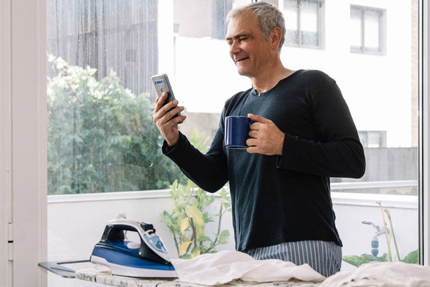 Volwassen man met een kopje koffie en kijken naar mobiele telefoon tijdens het strijken shirt thuis - Foto, afbeelding