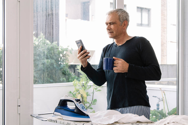 Mature man having cup of coffee and looking at mobile phone while ironing shirt at home - Foto, immagini