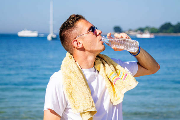 young man on the beach resting and drinking after exercising - Foto, imagen