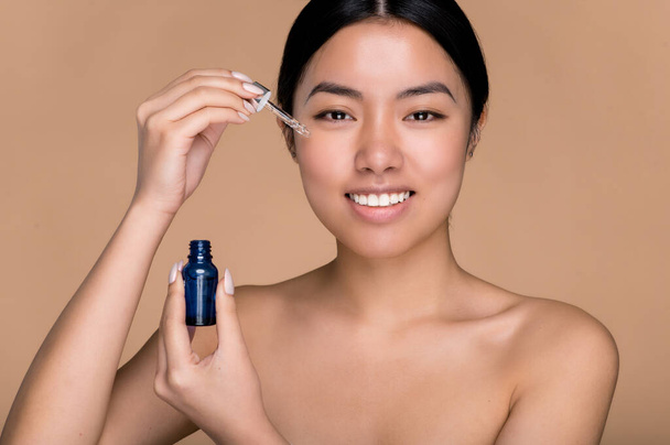Beauty and cosmetology concept. Young japanese woman with perfect skin without wrinkles applying hyaluronic serum on her face with pipette, looks at camera, smiles, stands on isolated beige background - 写真・画像