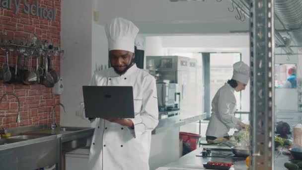 Afrykański amerykański kuchenka patrząc na ekranie laptopa gotować danie dla smakoszy - Materiał filmowy, wideo