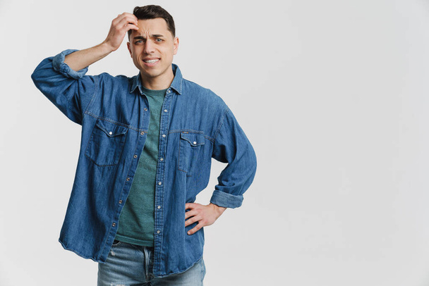 Young brunette man dressed denim shirt frowning and scratching his head isolated over white background - 写真・画像