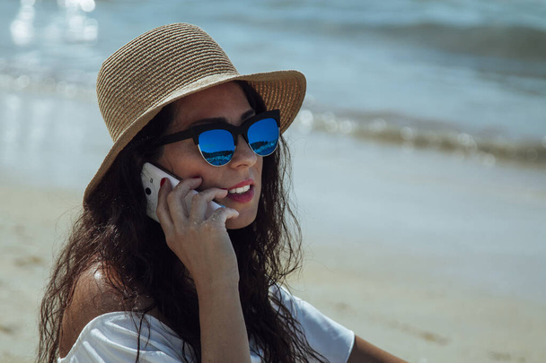 женщина в шляпе разговаривает с мобильным телефоном, сидя на пляже - Фото, изображение