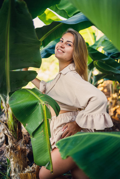 Woman near green leaves of banana bushes on nature in a park in a tropical place, she is in a beige skirt and a blouse - Foto, Imagem