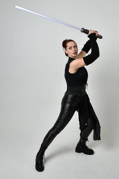 Портрет повної довжини досить червоного волосся жіночої моделі в чорному футуристичному шкіряному костюмі, що тримає зброю для меча з вогню. Динамічні стоячі пози з жестовими руками, звернені назад на білому фоні студії
. - Фото, зображення
