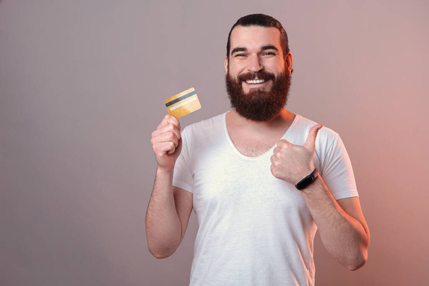Ευτυχισμένος νεαρός άνδρας με γενειάδα κρατώντας κίτρινη χρεωστική κάρτα και δείχνοντας τον αντίχειρα επάνω χειρονομία. - Φωτογραφία, εικόνα